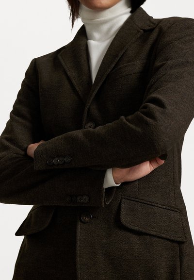 Dark brown blazer made of textured fabric, featuring notch lapels, two buttons, and pockets, layered over a white turtleneck.