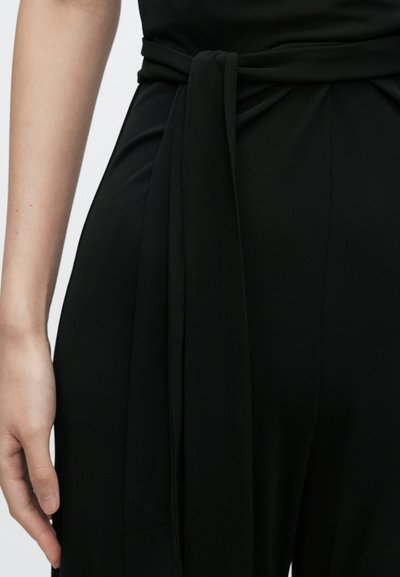 Črni kombinezon z detajlom zavezovanja v pasu, gladko teksturo tkanine in prilegajočo se silhueto, roka je položena ob strani.