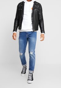 Černá kožená bunda, bílá mikina s oranžovým logem, světle modré roztrhané džíny a černé vysoké tenisky. Ležérní vrstvený outfit.