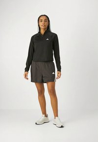 Camiseta deportiva negra de manga larga con media cremallera y pequeño logo, combinada con pantalones cortos a juego. Zapatillas deportivas blancas con suelas texturizadas.