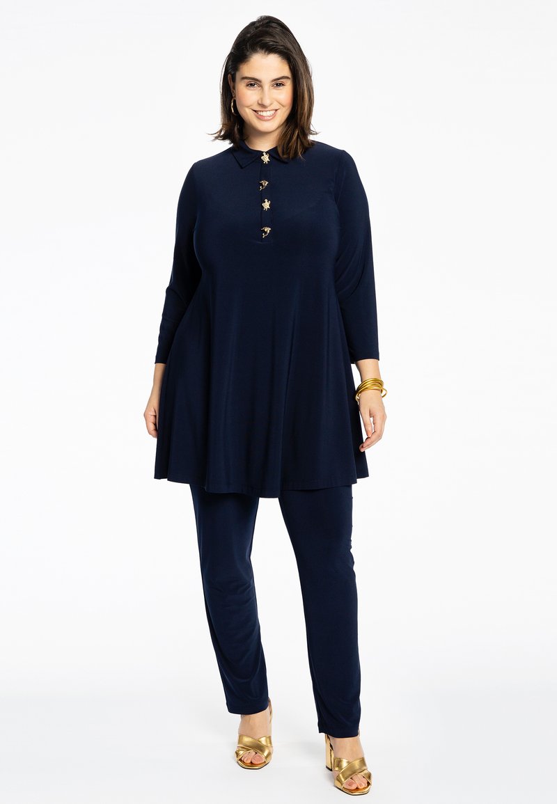 Marine tuniek met een kraag en gouden knoopaccenten, gecombineerd met bijpassende broeken. Glad materiaal, relaxte pasvorm en enkel-lang. Gouden sleehaksandalen.