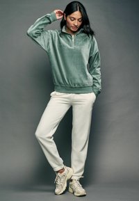 Grøn kordfløjl quarter-zip pullover med ribbestrikket krave, parret med hvide tapered joggers og beige løbesko.