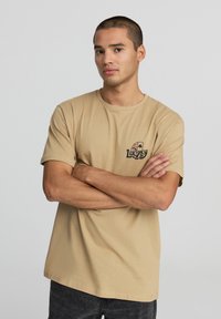T-shirt in cotone beige con logo sul petto che raffigura delle carte da gioco e la parola "Lucky". Maniche corte, scollo rotondo, orlo diritto.