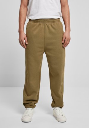 Mężczyzna ubrany w oliwkowozielone spodnie dresowe z elastycznym pasem i białe tenisówki, stojący na tle jednolitego jasnoszarego tła.