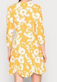 Robes fleuries sur un fond jaune avec des fleurs blanches. Elle a des manches longues et un ourlet froncé en bas.