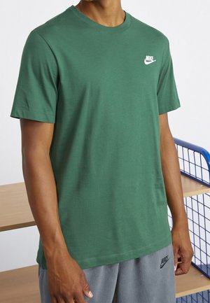 Man wearing green Nike t-shirt and gray Nike shorts standing near wooden shelves with blue metal frame.