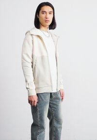 Lys beige zip-hette med hvit innvendig, med sidelommer og mansjetterte ermer, iført over en hvit t-skjorte og grå jeans.