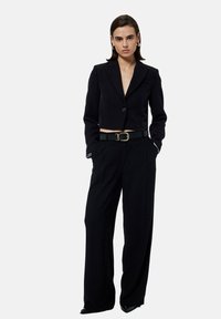 Zwarte cropped blazer met één knop, gecombineerd met zwarte hoge taillebroeken. Beschikt over een bijpassende zwarte riem met een gouden gesp.