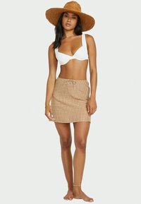 Bílý bikini top a hnědá kostkovaná mini sukně s tkaničkou. Klobouk se širokým okrajem z otavu. Bosá, s decentním šperkem na kotníku.