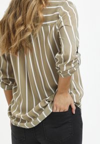 Une personne avec de longs cheveux ondulés porte une chemise rayée beige et blanche et un pantalon noir, la main placée dans la poche arrière, faisant face à l'arrière.