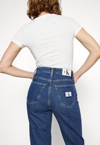 Siniset korkeavyötäröiset farkut suoralla leikkauksella, valmistettu denimi-kankaasta, joissa on kaksi takataskua ja merkkilaput vyötäröllä ja taskussa.