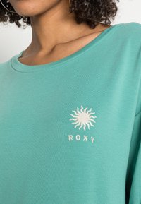 T-shirt di cotone turchese con scollatura rotonda e grafica di un sole con "ROXY" ricamato in bianco. Tessuto morbido, vestibilità rilassata.