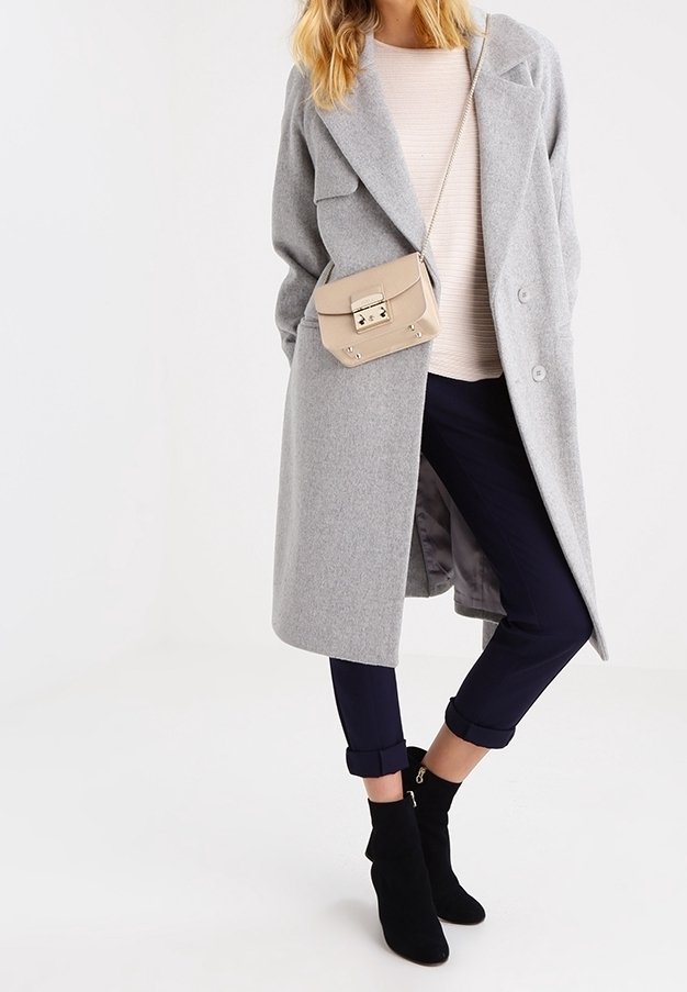 Cappotto di lana grigio con ampi revers, borsa a tracolla beige, maglione lavorato a maglia color crema, pantaloni cropped blu navy e stivaletti neri con cerniere.