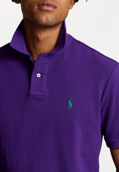 Polo shirt violet en tissu texturé, avec un col rabattu, une patte de boutonnage à deux boutons et un petit logo vert sur la poitrine.