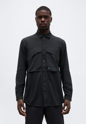 Mees, kellel on seljas must pikkade varrukatega nööbitav särk koos eest taskuga, seisab ühetaustalise heleda tausta ees.