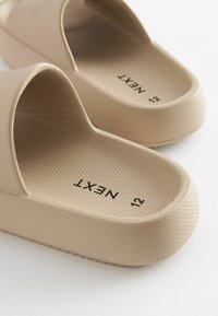 Beige formede klipklapper med tekstureret overflade og åbent tådesign. Interiøret har en grebtekstur; størrelse trykt som "12" indeni.