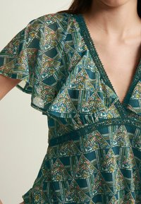 Bluse aus durchsichtigem, grünem Stoff mit Blumenmuster, Rüschärmel, V-Ausschnitt und Spitze entlang des Ausschnitts und der Taille. Sichtbare geometrische Designdetails.