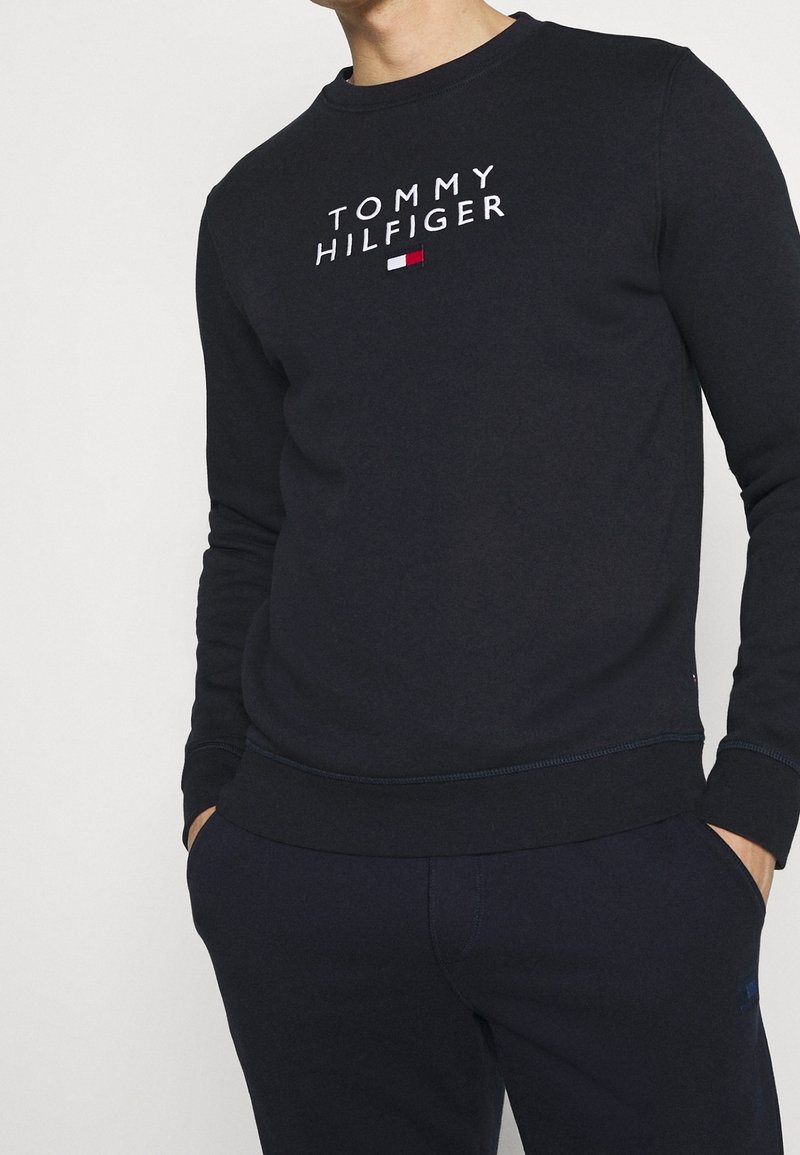 Homme portant un sweat-shirt bleu marine Tommy Hilfiger et un pantalon assorti, les mains dans les poches, visible du cou aux hanches.