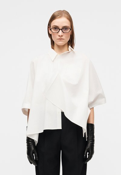 Weiße Baumwollbluse mit kurzen Ärmeln, asymmetrischem Saum, linker Brusttasche, getragen mit schwarzen Lederhandschuhen und dunklen Hosen.