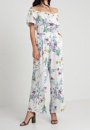 Kvinde iført hvid off-shoulder blomstrejumpsuit med vide ben og grønne hæle, stående imod en ensfarvet baggrund.