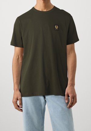 Man wearing olive green short-sleeve t-shirt with small black and yellow logo on chest, paired with light blue jeans.