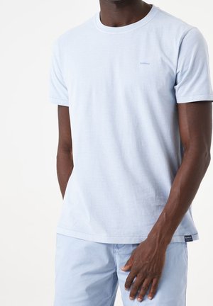Mann trägt hellblaues kurzärmeliges T-Shirt und passende Shorts, steht vor neutralem weißem Hintergrund.