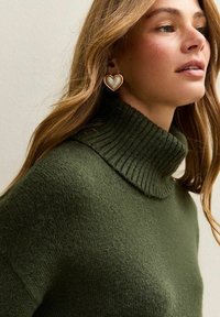 Groene trui met hoge nek, ribbelstructuur en een losse fit, gecombineerd met grote hartvormige oorbellen met een gouden rand en een witte center.