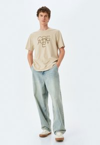 Beige bomulds T-shirt med "CREATIVE" trykt i brun, parret med lyseblå løstsiddende jeans og beige sneakers med brune detaljer.