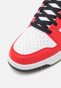 Zapatilla roja y blanca con parte superior de cuero, cordones negros, puntera perforada para transpirabilidad y suela de goma de contraste. Detalles texturizados visibles en los laterales.