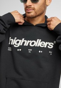 Fekete kapucnis pulóver, elől "highrollers" felirattal és játszikártya szimbólumokkal díszítve. Az anyag lágynek tűnik; elöl zsebbel és állítható kapucnival rendelkezik.