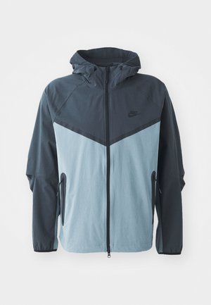 Windbreaker-Jacke mit Kapuze, vorderem Reißverschluss, dunkelblauem und hellblauem Blockdesign sowie seitlichen Reißverschlusstaschen.