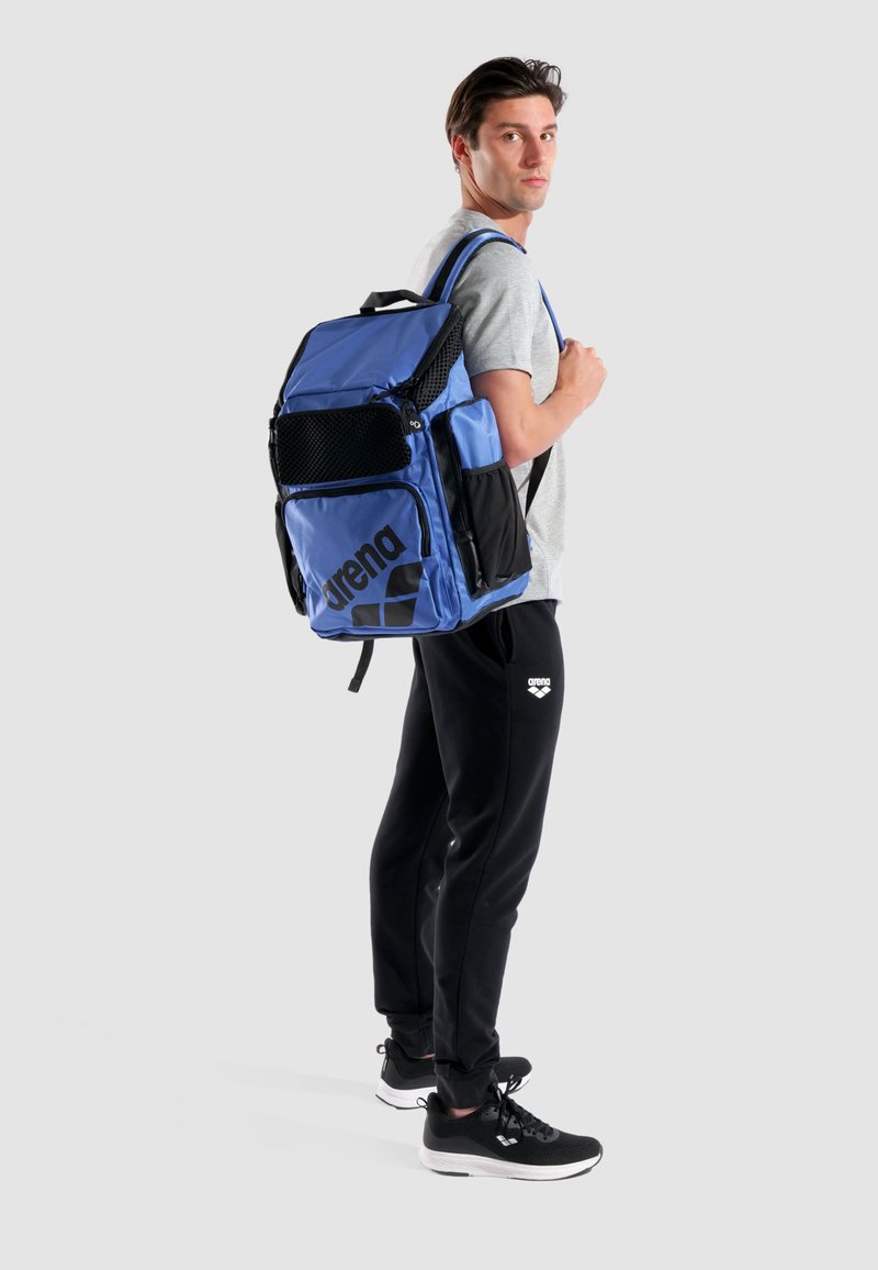 Mochila deportiva azul y negra con bolsillos de malla, múltiples cremalleras y un gran logo. Diseñada para la comodidad con correas acolchadas.