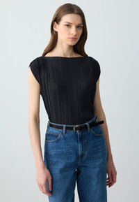 Top nero a pieghe con scollatura a barca, abbinato a jeans di denim blu a vita alta e una cintura nera. Tessuto liscio, design aderente.