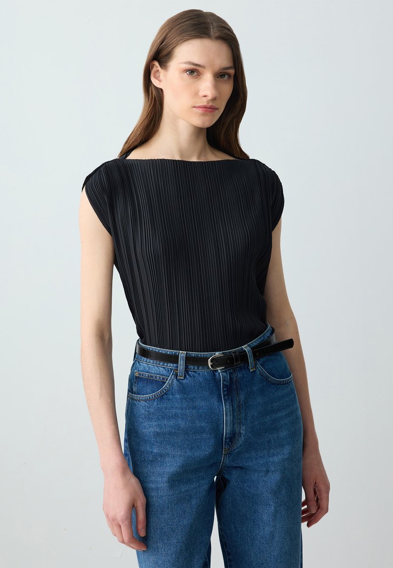 Top nero a pieghe con scollatura a barca, abbinato a jeans di denim blu a vita alta e una cintura nera. Tessuto liscio, design aderente.