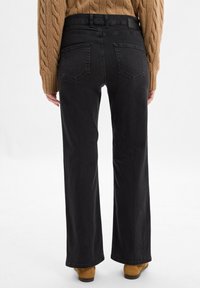 Schwarze, weit geschnittene Jeans mit hoher Taille, zwei Gesäßtaschen und einer glatten Textur. Kombiniert mit einem hellbraunen Grobstrickpullover und braunem Schuhwerk.