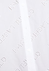 Chemise blanche boutonnée avec détails en texte découpé, texture lisse et petites fermetures à boutons sur le devant.