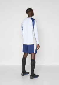Completo sportivo composto da una maglietta bianca a maniche lunghe, pantaloni corti blu con dettagli beige, calze nere fino al ginocchio e scarpe da calcio nere con dettagli blu.