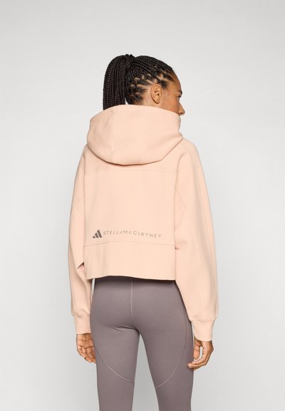 Skraćeni hoodie u mekoj ružičastoj tkanini s kapuljačom, koji ima logo na leđima i široke rukave, uparen s oblikovanim sivim tajicama.