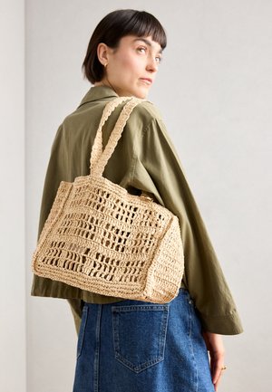 Sac en crochet tissé de couleur beige, de forme rectangulaire, avec deux larges poignées et un motif en maille ouverte. Le design présente un tissu texturé et un intérieur spacieux.