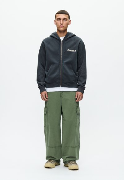 Junger Mann steht, trägt einen schwarzen Kapuzenpullover mit Reißverschluss und "Double V" Logo, grüne Cargo-Hose, beige Turnschuhe und ein weißes Hemd darunter.