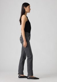 Zwarte mouwloze top gecombineerd met hoge grijze jeans met een ontwerp voor een achterzak en zwarte platte schoenen. Neutrale achtergrond.