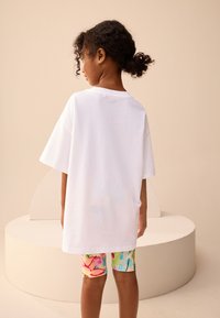 Witte katoenen t-shirt in oversized model met korte mouwen, gecombineerd met kleurrijke fietsbroek met bloemmotief. Effen achtergrond met zachte rondingen.