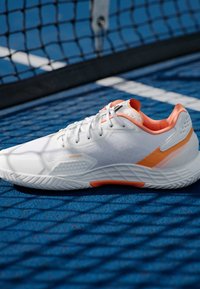 Λευκό αθλητικό παπούτσι με πορτοκαλί λεπτομέρειες, πλέγμα επάνω μέρος, υφή σόλας και κούμπωμα με κορδόνια, σε φόντο μπλε γηπεδική με δίκτυ.