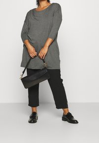 Haut gris à manches longues avec un décolleté en scoop; associé à un pantalon noir et un sac bandoulière noir. Le haut est doux, tombe délicatement et présente des détails sur les côtés.