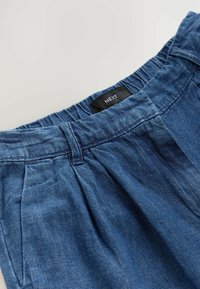 Denim püksid elastse vöökoha ja ees pleatidega. Värv on kesk-sinine ja tekstuur pehme. Brändi silt näitab "NEXT".