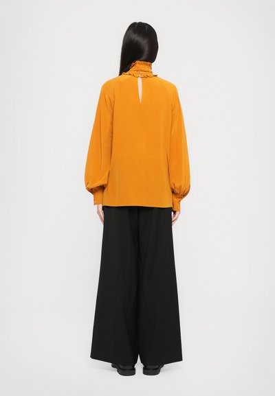 Mosterdgele blouse met een rimpelband en een keyhole achterkant, gecombineerd met brede zwarte broeken. Soepele stof en een ontspannen pasvorm zijn duidelijk zichtbaar.