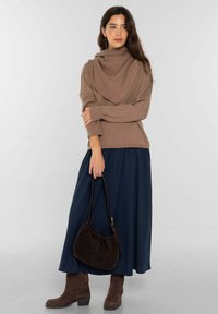 Suéter de punto marrón con cuello alto drapeado, combinado con una falda maxi plisada azul marino. La modelo sostiene un bolso de ante marrón oscuro.