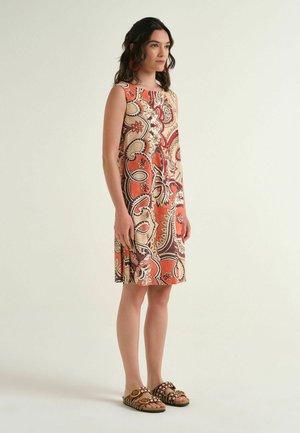 Donna in piedi che indossa un vestito senza maniche al ginocchio con motivo paisley rosso, beige e marrone e sandali slip-on con dettagli a fibbia.