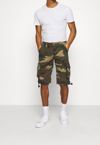 Pantaloni cargo mimetici con due tasche laterali e coulisse all'orlo, realizzati in tessuto misto cotone; abbinati a una t-shirt aderente bianca e sneakers bianche.