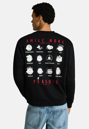 Mężczyzna widziany od tyłu, ubrany w czarną bluzę z czerwonym napisem "Smile More Worry Less" oraz białe postacie i nazwy Peanuts.